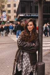 Obraz premium Portrait of a confident stylish woman with long voluminous hair and a calm facial expression looking at camera, leaning on a pillar on a busy city street background wearing a vintage leather jacket