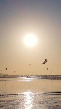Kitesurfing at sunset over the ocean with golden light reflecting on wet sand, multiple kites in the sky, and a tropical beach scene