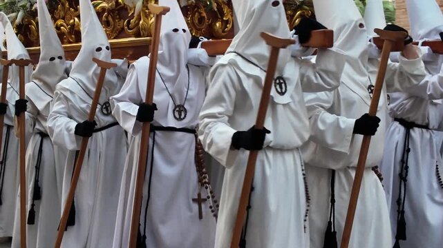 White robed nazareno penitents carrying wooden staffs during traditional Spanish Holy Week procession in Semana Santa religious street celebration