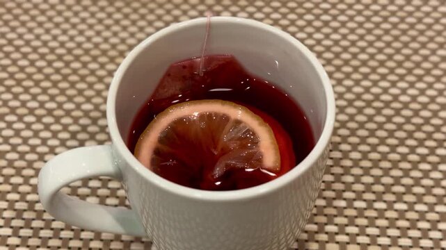 Close-up red karkade tea in cup lemon slice steeping porcelain mug on beige woven placemat, warm steam rising, tea bag string visible.