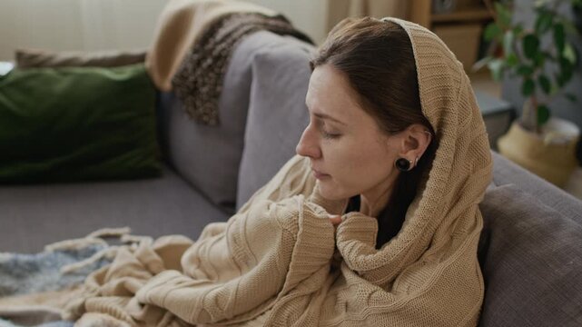 Medium shot of young Caucasian girl sitting on couch, wrapped in blankets, coughing and covering mouth with tissue, while recuperating on sick leave with flu, pneumonia or covid alone at home