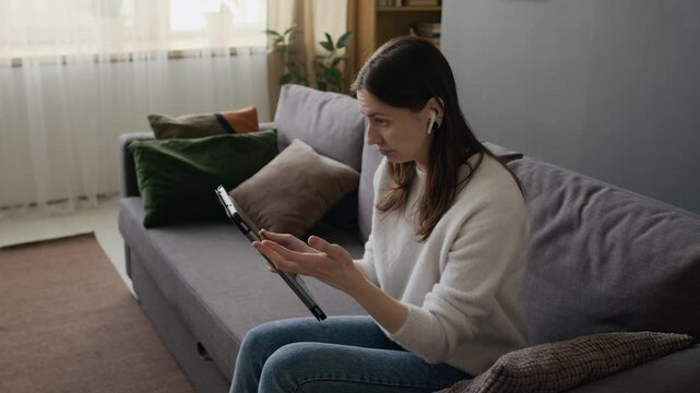 Medium shot of dismayed Caucasian girl sitting on couch at home, trying to connect to internet with digital tablet computer, experiencing wifi problems or mobile network outage