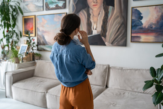 Thoughtful concentrated woman artist looking at paintings hanging on wall at home loft gallery, thinking getting inspiration for future artworks, enjoying skills progress and creativity development.