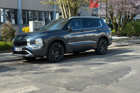 Modern dark grey Mitsubishi Outlander PHEV parked on city street, plug-in hybrid electric vehicle with black alloy wheels and EV badge, hybrid SUV, urban eco-friendly family transportation Frankfurt