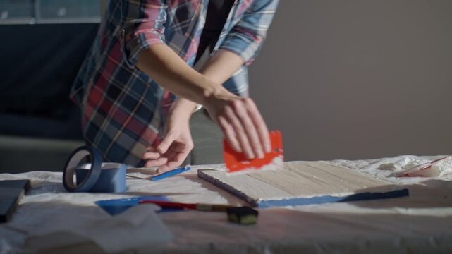 Woman spreading textured paste on white canvas using scraper on work table with art tools