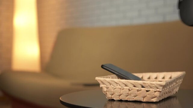 Man practicing digital detox reading book at home. Relaxed man putting his phone and headphones away in a basket on the coffee table, sitting on the sofa, and reading a book in his cozy living room