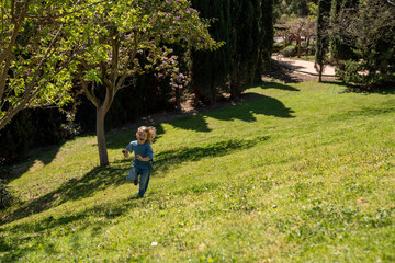 Obraz premium Little blond boy running on green hill in sunny park, childhood freedom concept, outdoor activity, joyful movement, spring nature, lifestyle scene, carefree play, landscape background