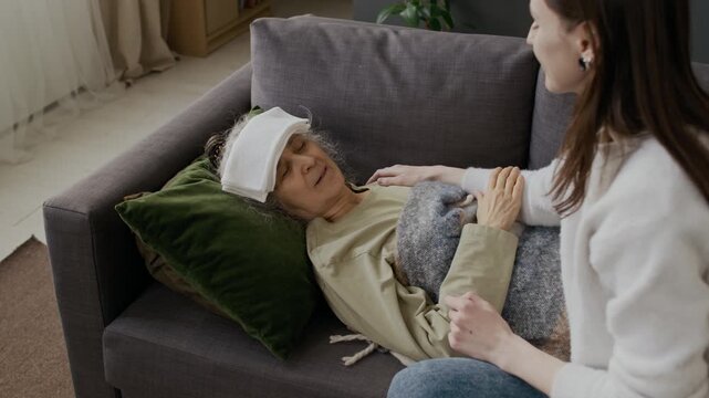 Medium shot of young Caucasian granddaughter putting cold towel compress on forehead for grandmother sick with flu, lying on couch with eyes closed, touching hand with gratitude and love