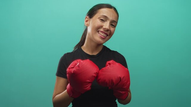 Young hispanic woman with red boxing gloves holding fists at chest in turquoise studio; confidence joy fitness.