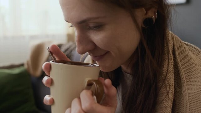 Close-up of face of smiling young Caucasian woman smelling hot tea with honey, then sipping to relieve sore throat pain, while recovering from seasonal cold or flu at home