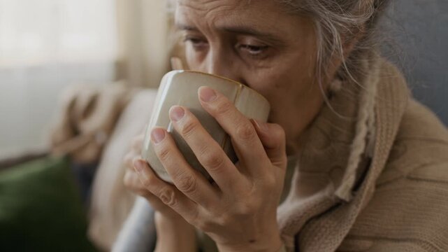 Close-up of face of senior Caucasian woman, sick with seasonal flu, drinking hot herbal or lemon and honey tea to recover from infection, feeling relief, blanket wrapped around shoulders