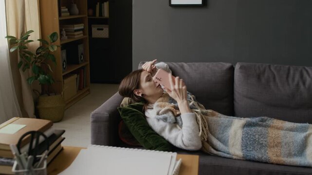 Full shot of young Caucasian woman lying on couch at home, covered with blanket, coughing with labored breathing and chest pain, then calling ambulance on mobile phone for urgent medical help
