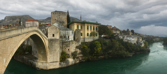 Naklejka premium Stari Most bridge in Mostar, Bosnia and Herzegovina, spanning the Neretva River under a dramatic cloudy sky with historic architecture and lush surroundings. 