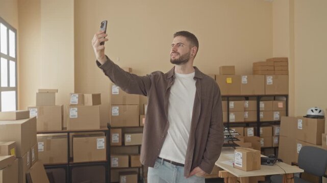 Man holds smartphone at arm's length with right hand, smiling amid stacked parcels in a packing building; entrepreneurial optimism.