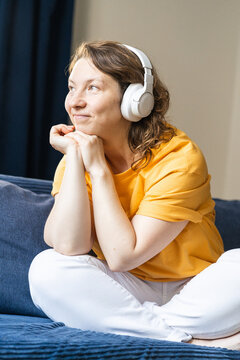 Woman in a yellow shirt and white pants sitting comfortably on a sofa, smiling while listening to music with headphones. Young happy woman enjoying music