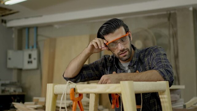 Carpenter woodworking workshop craftsman man shaping wood for handmade furniture indoor craft studio, measuring and clamping frame with tool workbench, wearing safety glasses, sanding furniture frame