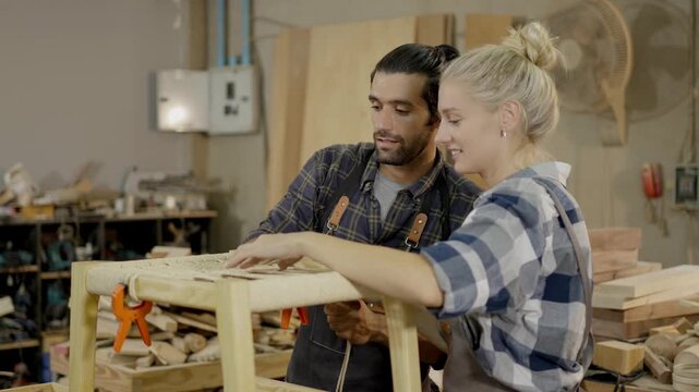 Carpenter woodworking workshop furniture table teamwork wood craftsman craftswoman tool two carpenters and apprentice sanding measuring clamping wooden table and stool studio, focused guidance
