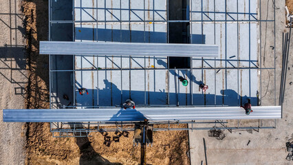 Aerial view of a yellow excavator lifting metal roofing sheets onto a steel frame. Construction workers installing a warehouse roof on a commercial building site with concrete foundation. © ultramansk