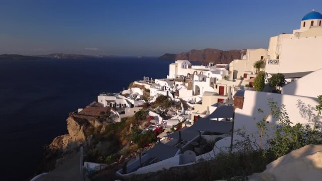 Picturesque whitewashed buildings cascade down volcanic cliffs in Oia village on Santorini island. Blue dome church and terraces overlook the deep blue Aegean Sea with distant caldera views. Greece