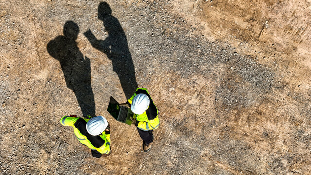 Aerial top down view of two engineers in safety gear discussing a project on a digital tablet. Construction workers standing on a dirt field casting long dark shadows in the bright sunlight.