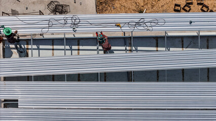 Naklejka premium Aerial view of construction workers installing roofing panels on a building, surrounded by tools and materials, showcasing teamwork, precision, and the construction process in progress.