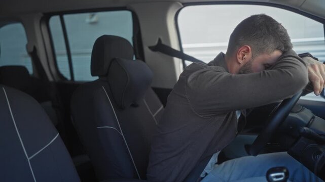 Young hispanic man in car wearing casual jacket and seatbelt, hands on steering wheel rubbing his eyes with a short beard, seated inside a parked vehicle on a street; fatigue reflection.