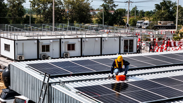 A worker wearing safety gear installing solar panels on a rooftop with nearby housing units in the background, emphasizing renewable energy, sustainability, and efficient solar power implementation
