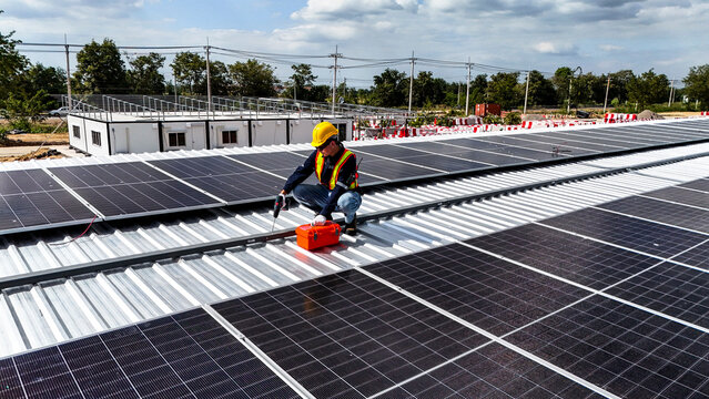 Top view of a solar power plant installation on a warehouse roof. Worker inspecting renewable energy modules. Sustainable architecture concept for eco friendly commercial infrastructure.