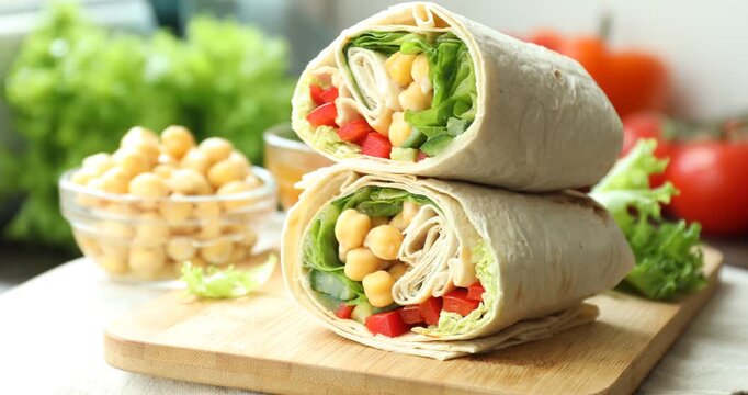 Board with pieces of tasty wrap with chickpeas, sauce and vegetables turning against blurred background, closeup