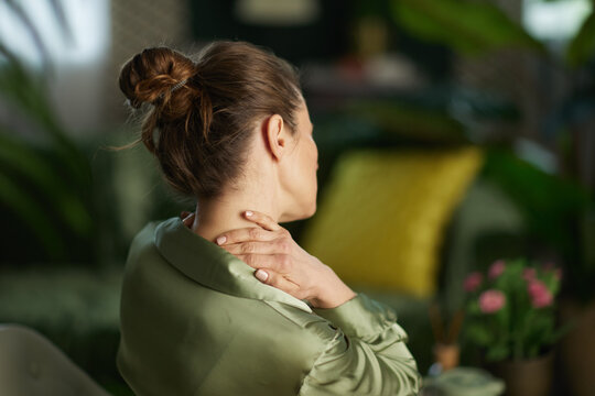 A woman in a green blouse reaches back to touch her neck, depicting muscle tension and the need for a mindful pause and physical relief in her home environment.