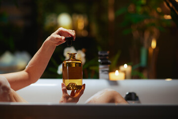 A person relaxes in a white bathtub, carefully opening a large golden bottle, creating a luxurious spa-like moment. Soft candle glow and blurred green foliage set a serene and indulgent mood. © Alliance