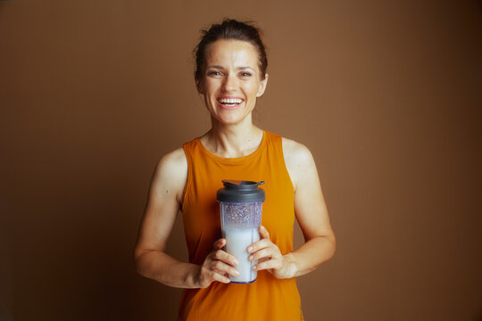 A smiling woman in a mustard-colored tank top holds a clear shaker bottle with a white liquid, embodying health, fitness, nutrition, and an active lifestyle.