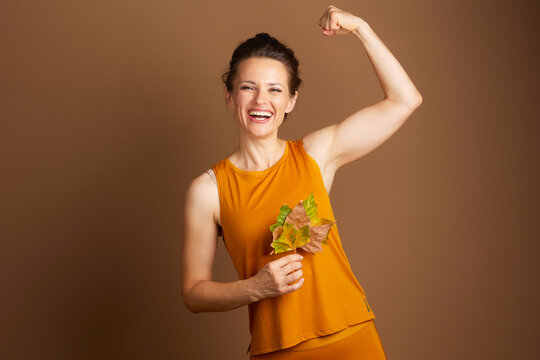 An adult woman in mustard yellow activewear laughs and flexes her bicep while holding autumn leaves, embodying strength, vitality, and cheerful seasonal health.