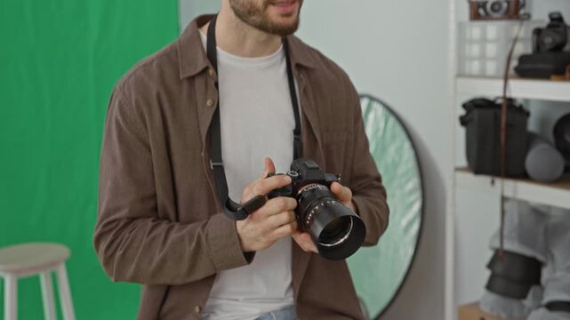Young man adjusts camera lens with face visible and holding camera strap in studio near green screen and reflector; creative concentration.
