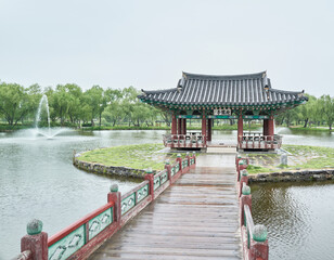 Fototapeta premium This serene photo captures a traditional Korean pavilion at Gungnamji Pond in Buyeo on a rainy day. A wet wooden bridge leads to the historic structure, surrounded by lush trees and calm waters.