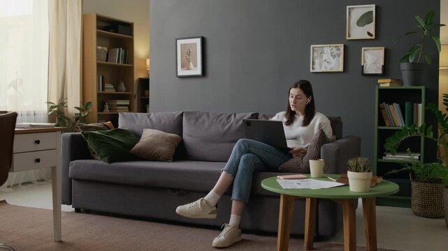 Wide shot of cheerful young Caucasian woman with earbuds sitting on couch in cozy living room at home, having animated chat with friend on video call on laptop, gesticulating and sharing life events