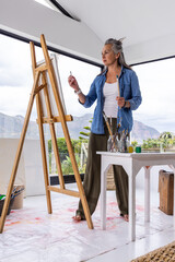 Naklejka premium Senior female artist standing barefoot in bright studio, painting on wooden easel above drop cloth
