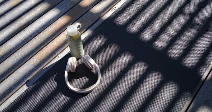 Pale bottle with loop cap, headphones sitting on dark mat on wooden deck, casting slatted shadows