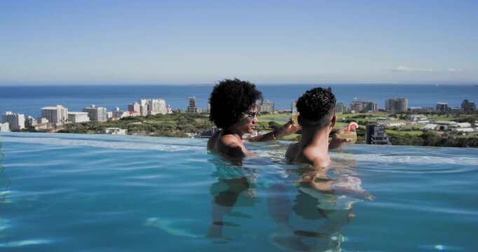 Diverse male friends wading in rooftop infinity pool, one raising citrus drinks, toasting, relaxing