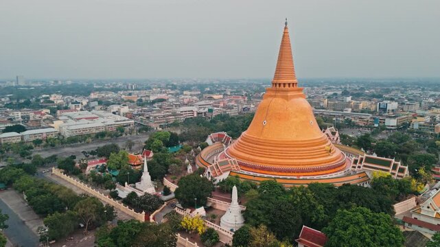 Camera moves in reverse along paved walkway revealing wide entrance leading toward giant orange stupa while hazy morning light illuminates green trees surrounding sacred Buddhist site in Thailand