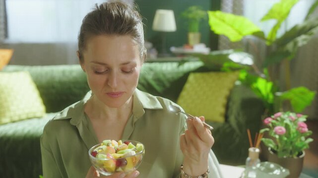 A middle-aged woman enjoys a bowl of colorful fruit salad, smiling at the camera in a bright, modern living room, symbolizing wellness and a balanced diet.