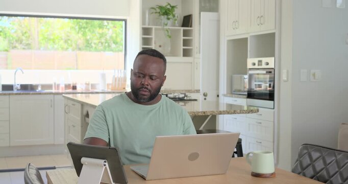 African man looking at laptop typing while checking tablet, mug at table working from home