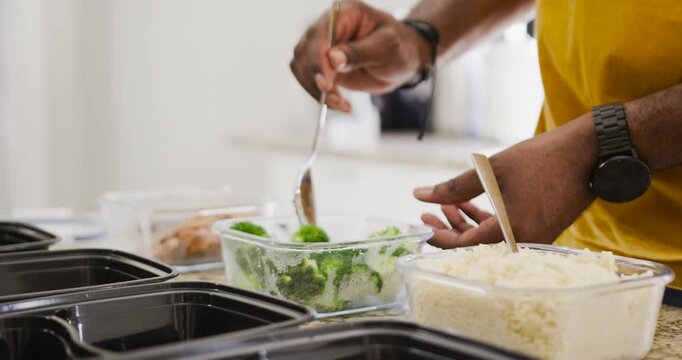 Post-cooking, man in yellow shirt scooping broccoli into meal-prep trays on counter, copy space
