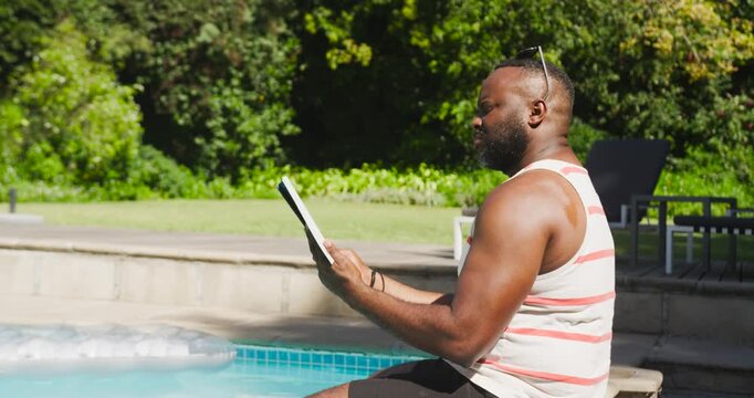 African American man sitting poolside reading book, flipping pages and adjusting shades, copy space