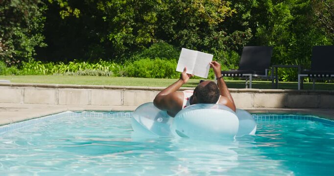 African man in red reclining on white ring in home pool, adjusting paperback against sun glare