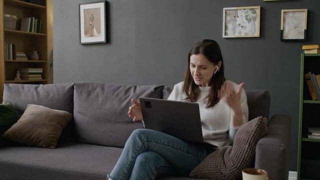 Medium full shot of frustrated young Caucasian woman with earphones sitting on couch at home, chatting on video call, losing internet connection, shaking laptop, swearing and putting aside in dismay
