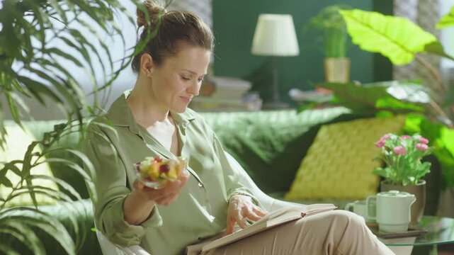 A woman sits comfortably in a sunlit living room filled with plants, reading a book and eating fresh fruit, showcasing a calm and healthy daily routine.