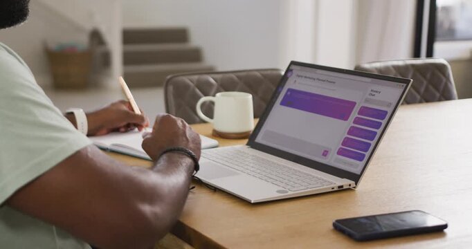 African American man referencing purple laptop app and writing notes in notebook at home