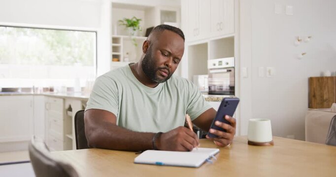 African man reading smartphone while copying into spiral notebook at kitchen table for home tasks