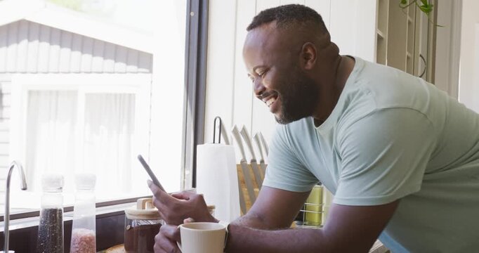 Smartphone buzzing African American midadult man in tee leaning on kitchen counter with mug reading